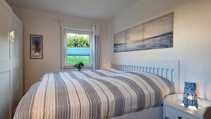 Un dormitorio con cama, una ventana con persianas, una mesita de noche con lámpara y un cuadro en la pared.