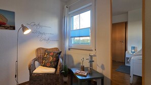 Una habitación acogedora con una silla de mimbre, una mesita con lámpara, una ventana con persianas y un cuadro en la pared.
