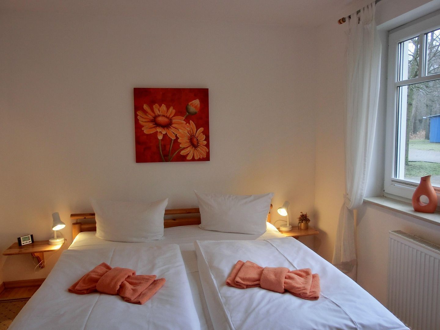 Habitación de hotel con dos camas, ropa de cama blanca, un cuadro de flores en la pared, una ventana con vistas al exterior y una mesita de noche con una lámpara y un pequeño jarrón.