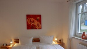 Una camera d'hotel con due letti, lenzuola bianche, un quadro con fiori sulla parete, una finestra che offre vista sull'esterno e un comodino con una lampada e un piccolo vaso.