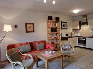 A cozy living room with a red sofa, a wooden coffee table, and wicker chairs. A bookshelf with books and speakers is in the background.
