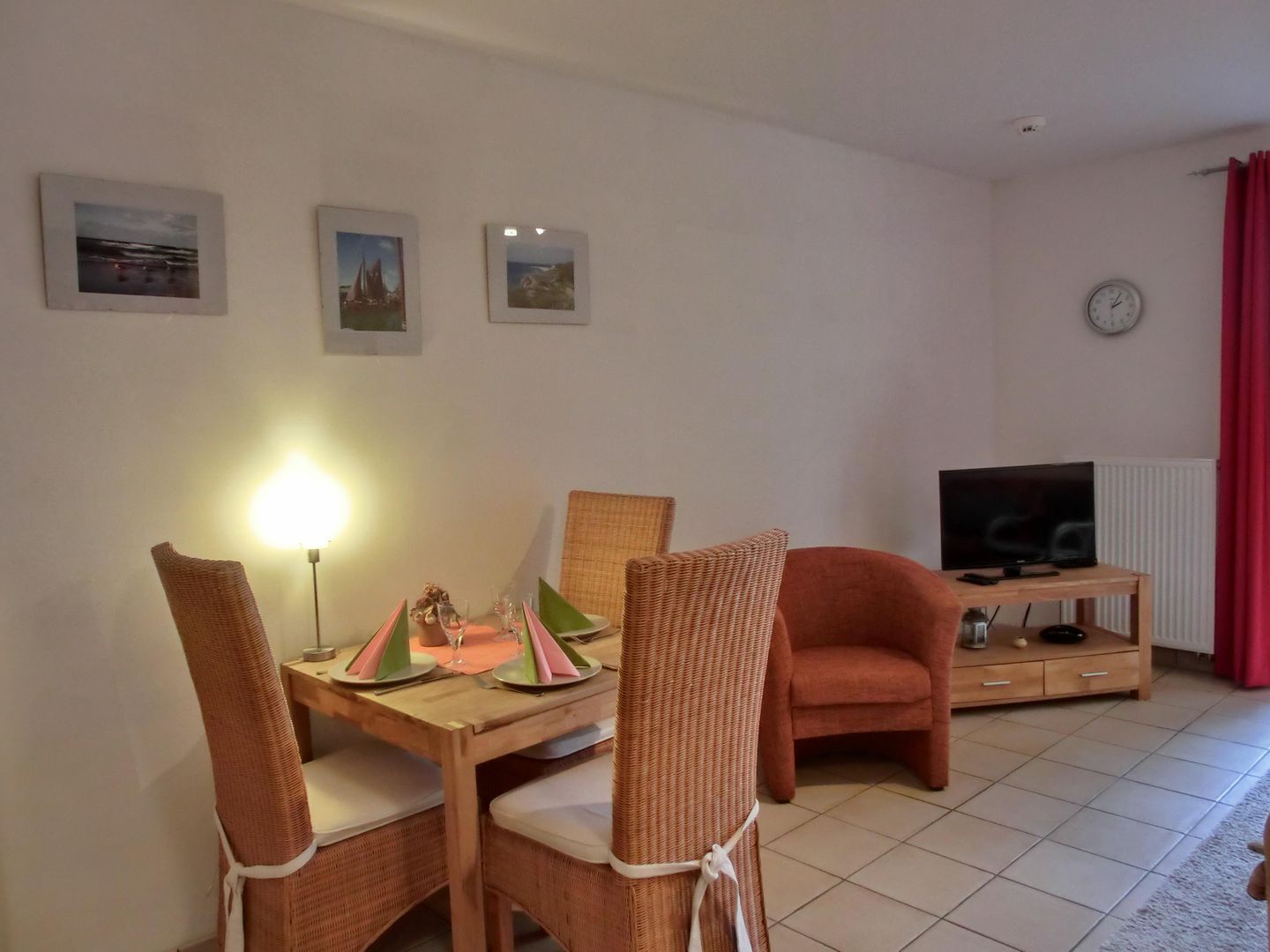 A dining area with wicker chairs and a table set for two, a television on a wooden stand, and a clock on the wall.