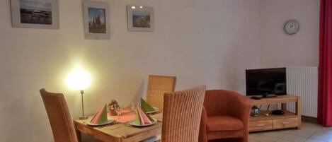 A dining area with wicker chairs and a table set for two, a television on a wooden stand, and a clock on the wall.