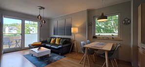 A modern living room with a black leather sofa, a white coffee table, a dining table with chairs, and a large window with a view of greenery.
