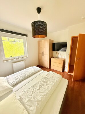 Une chambre avec deux lits, une armoire, une commode, une télévision et une fenêtre donnant sur des arbres.