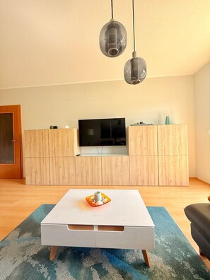 A modern living room with a flat-screen TV, wooden cabinets, a white coffee table, and two hanging pendant lights.