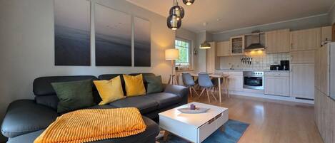 A modern living room with a black sofa, yellow cushions, and a white coffee table. There is a kitchen area with wooden cabinets and a stove.