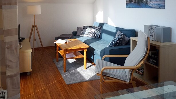 A modern living room with a wooden floor, a blue sofa, a wooden coffee table, a glass table with items, and a painting of a beach scene on the wall.