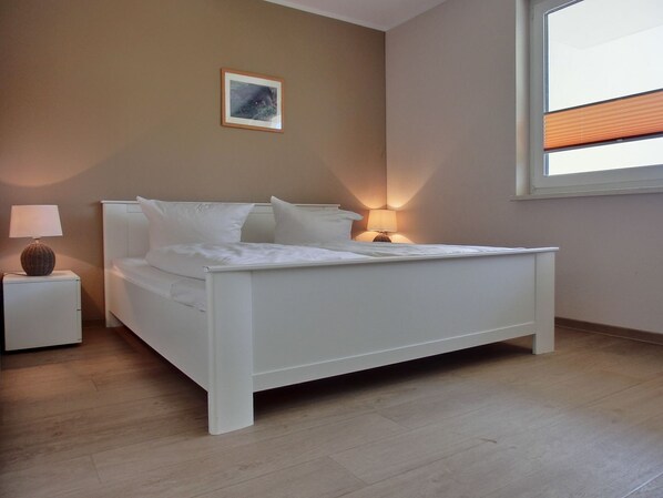 Une chambre avec un lit blanc, une table de chevet avec une lampe, un tableau encadré au mur et une fenêtre avec des stores.