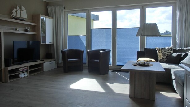 Sala de estar moderna com televisão de tela plana, duas poltronas, um sofá, uma mesa de centro e uma janela grande com vista para o mar.