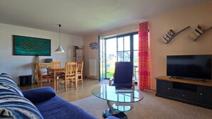 Un soggiorno con divano, tavolino in vetro, televisore su un mobile, zona pranzo con sedie e tavolo, e una grande finestra con tende.