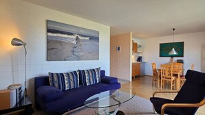 Un soggiorno con un divano, un tavolino in vetro, un quadro raffigurante una spiaggia, una zona pranzo con tavolo e sedie, e una cucina sullo sfondo.