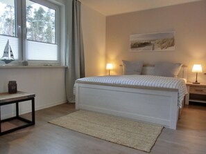 A bedroom with a bed, two bedside tables with lamps, a window with curtains, a small table with a jar, and a rug.