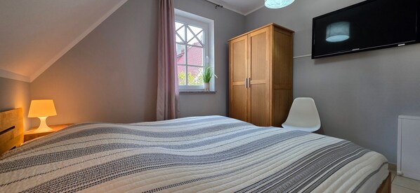 Une chambre à coucher comprenant un lit, une table de chevet, une lampe, une armoire, une chaise, un téléviseur et une fenêtre avec des rideaux.