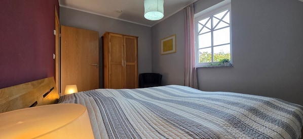 A bedroom with a bed, wooden wardrobe, a window with a view, and a wall-mounted picture.