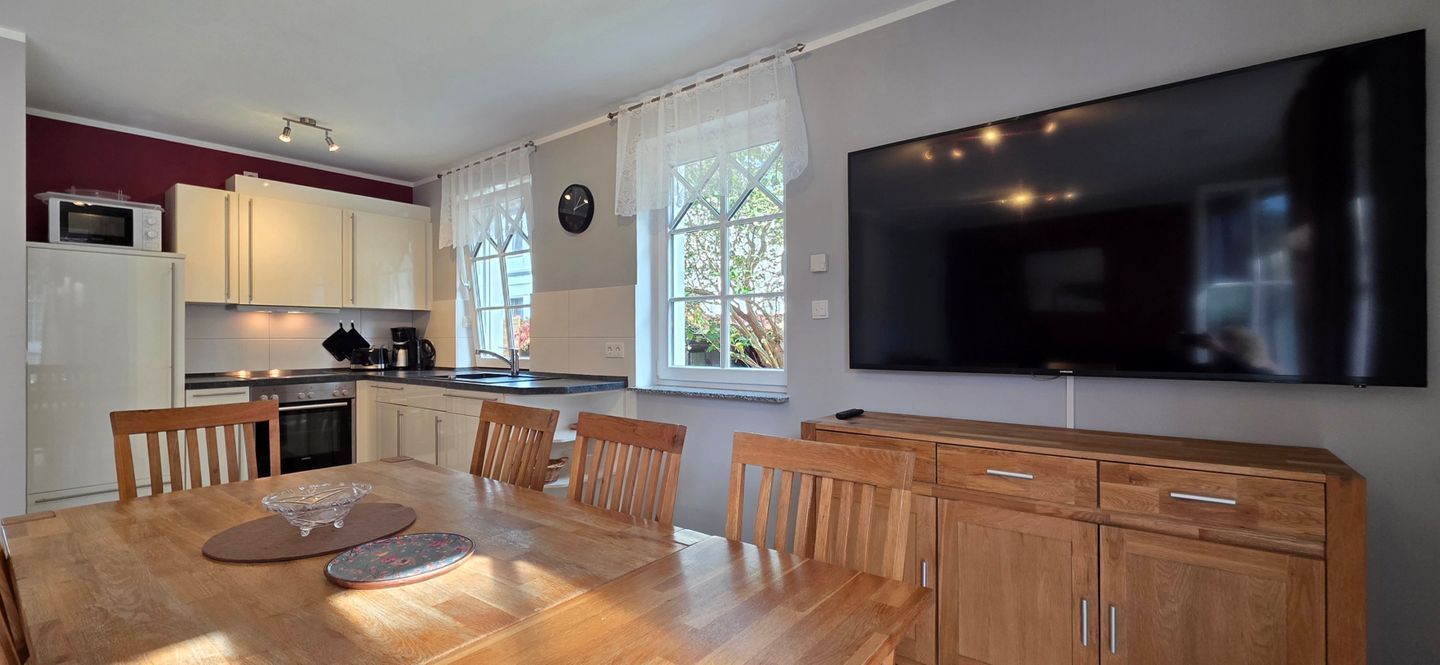 A modern kitchen with white cabinets, a wooden dining table, and a flat-screen TV mounted on the wall.