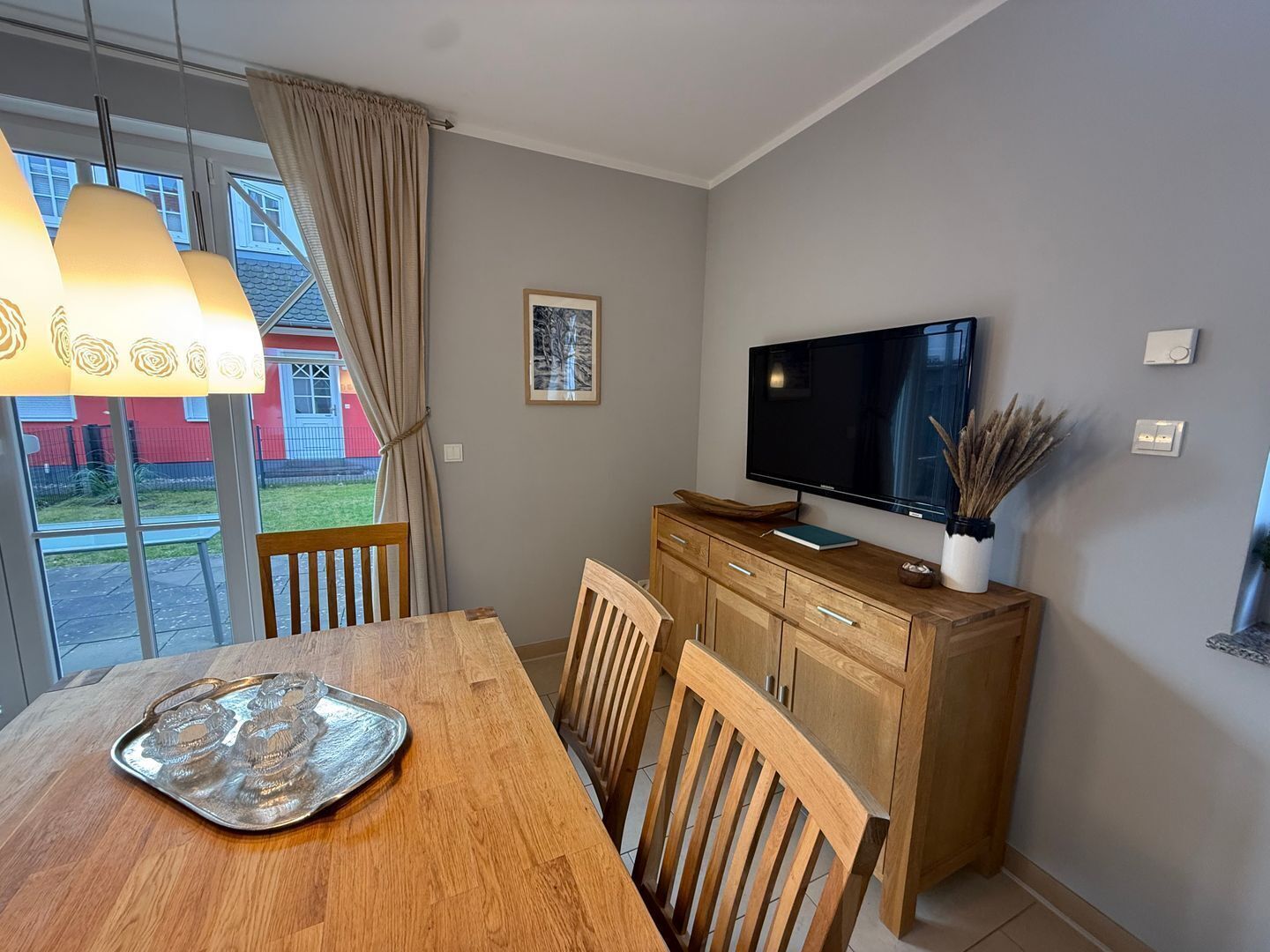 Un coin repas avec une table en bois, des chaises, un téléviseur posé sur un meuble en bois et une fenêtre avec des rideaux.