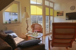 A cozy living room with a sofa, a wooden chair, a table with a lamp, and a view through large windows.