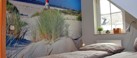 A bedroom with a large beach mural, a window with a view of the sea, and a bedside lamp.