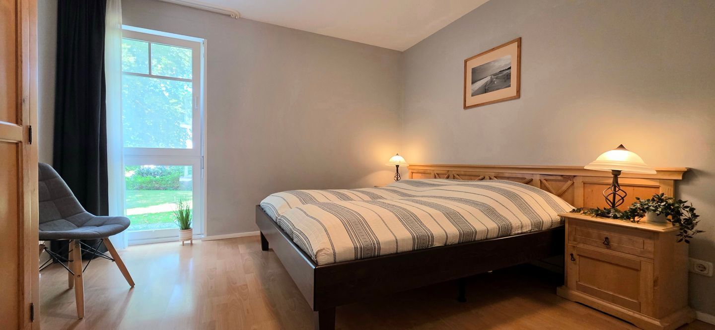 A bedroom with a bed, bedside table, lamp, chair, and a window with a view of greenery.