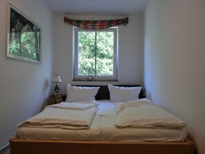Un dormitorio con cama, ventana con vista a árboles, lámpara y un cuadro en la pared.