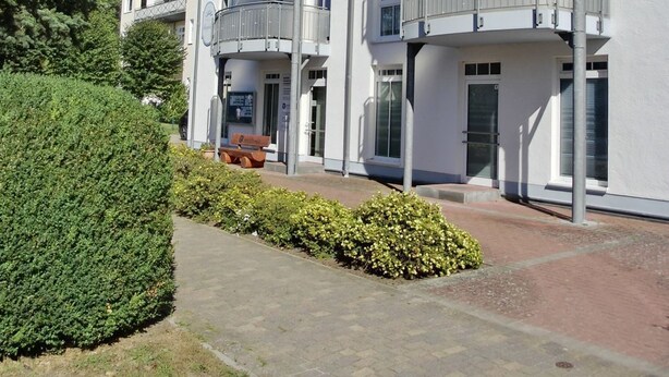 A multi-story residential building with balconies, a paved entrance, and a well-maintained hedge.