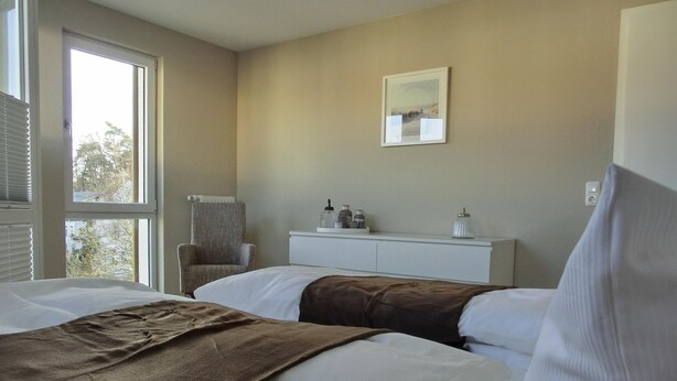 Une chambre d’hôtel avec deux lits, une table de chevet blanche, une chaise et une fenêtre avec des stores.