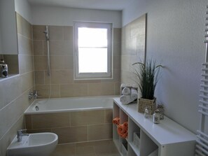 A modern bathroom with a bathtub, a shower, and a sink. There is a window providing natural light, and a shelf with towels and a plant.