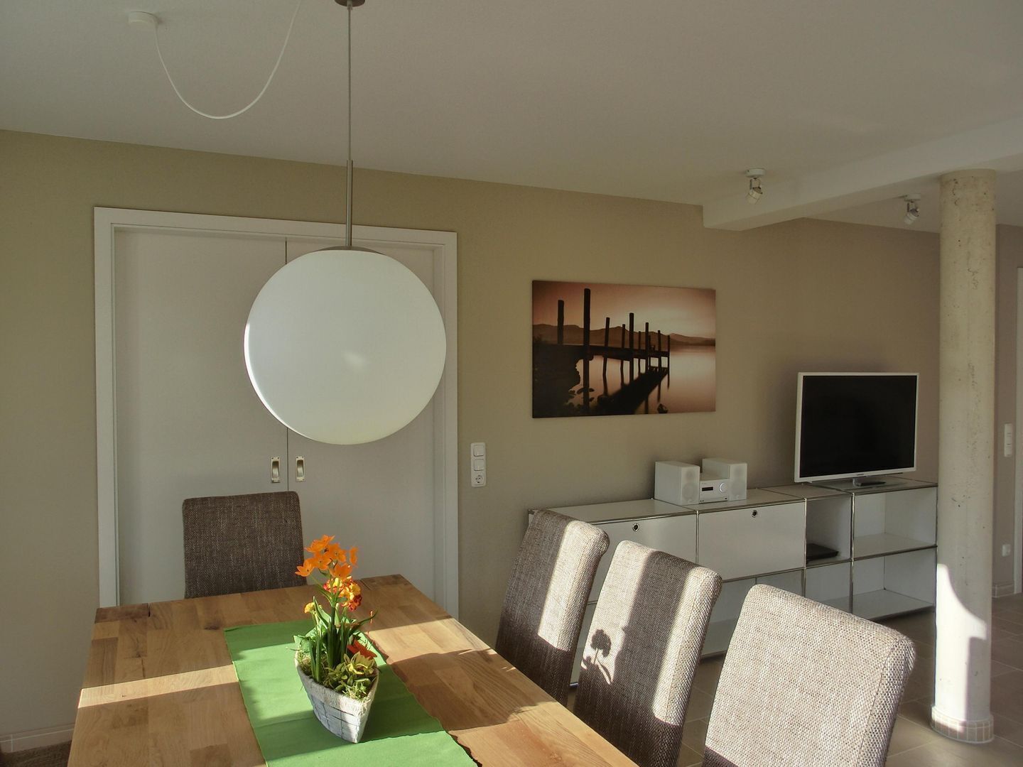 A dining area with a wooden table, chairs, a hanging light fixture, a painting, and a television on a stand.