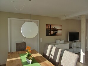 Uma sala de jantar com mesa de madeira, cadeiras, luminária pendente, quadro e televisão sobre um suporte.