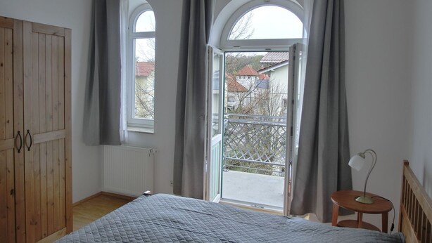 A bedroom with a bed, a wooden wardrobe, a small table with a lamp, and a balcony with a view of buildings. - Vacation apartment LVC4 - 2-room apartment for 4 pers. with WLAN (Graal-Müritz)