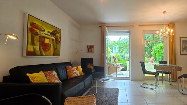 Uma sala de estar moderna com um sofá preto, uma mesa de jantar e cadeiras. Há um quadro grande na parede, um lustre e uma vista através da porta de vidro.