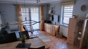 A modern living room with a dining area, featuring a sofa, a wooden dining table, and a TV mounted on the wall. - Vacation Rental K36 - Central 2-Bedroom Apartment for 3 People, Terrace (Graal-Müritz)