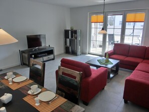 Een woonkamer met een rode bank, een eettafel gedekt voor twee personen, een televisie en een boekenkast.