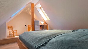 A bedroom with a sloped ceiling, a skylight, a bed with a blue blanket, a wooden chair, and a small desk with a lamp.