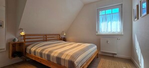 Une chambre avec un plafond en pente, un cadre de lit en bois, un radiateur et une fenêtre avec des rideaux.