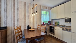A compact kitchen with white cabinets, a wooden dining table, and a hanging light fixture. - Apartment K21 "Storchennest" - 3-room apartment for 6 pers. on the 2nd floor with balcony (Graal-Müritz)