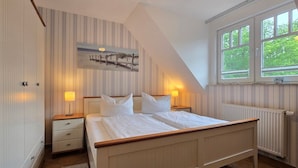 A bedroom with a bed, two bedside tables with lamps, a radiator, and a window with a view of trees. - Apartment K21 "Storchennest" - 3-room apartment for 6 pers. on the 2nd floor with balcony (Graal-Müritz)