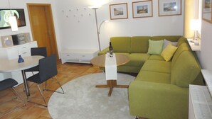 Sala de estar con sofá verde, una mesa redonda de centro, una mesita auxiliar blanca, televisor, lámpara y una pequeña mesa con una botella y vasos.