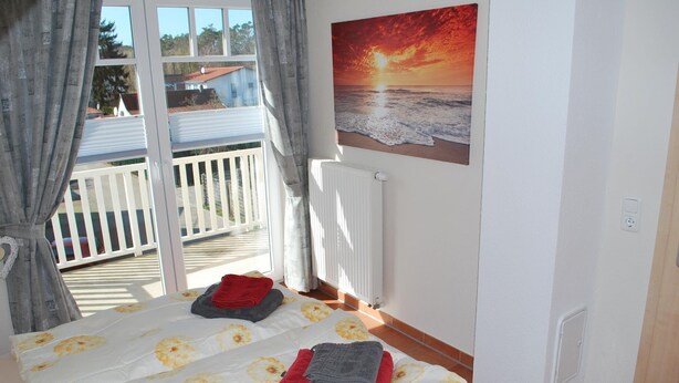 A bedroom with a bed, a window with curtains, a radiator, and a painting of a sunset. - Vacation apartment K38 "Sonnenblume" - 3-room apartment with 2 floors for a maximum of 6 people. (Graal-Müritz)