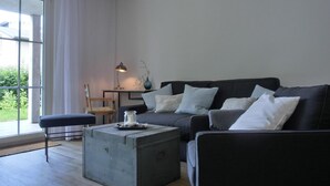 A modern living room with a dark grey sofa, a small wooden table, and a grey ottoman. A window with white curtains allows natural light into the room.