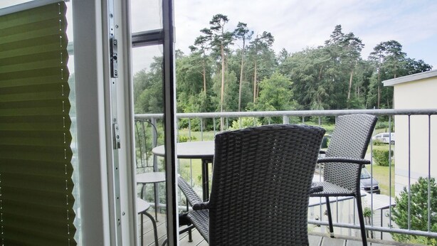 Un balcon avec des fauteuils et une table en osier, donnant sur un jardin planté de grands arbres.