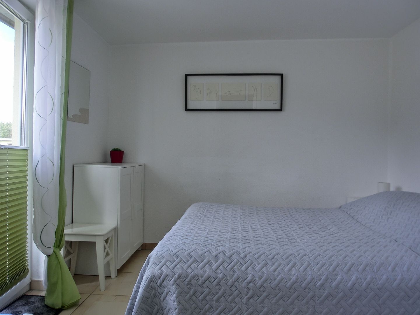 Une chambre à coucher avec un lit, une chaise, une petite table sur laquelle se trouve un objet rouge, un tableau encadré au mur et une fenêtre avec des stores.