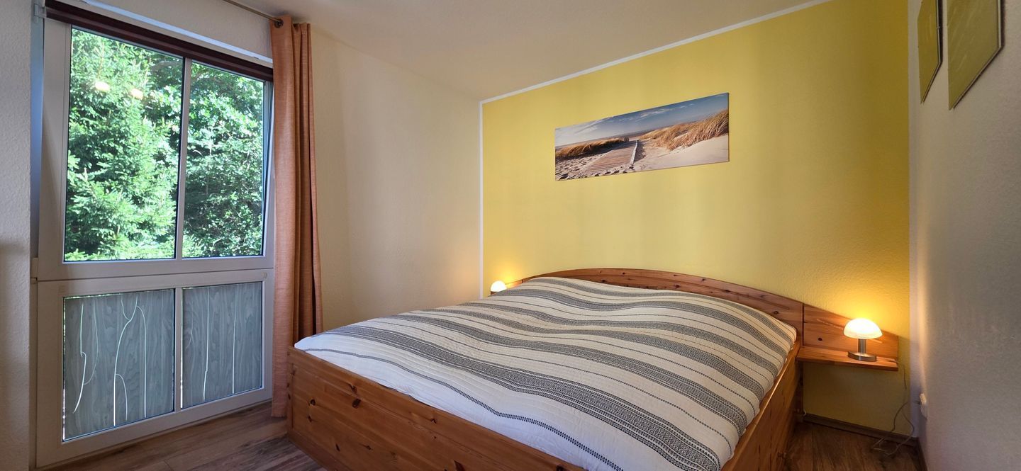 Ein Schlafzimmer mit einem hölzernen Bett, einem Fenster mit Vorhängen, einem an der Wand befestigten Bild und Nachttischlampen.