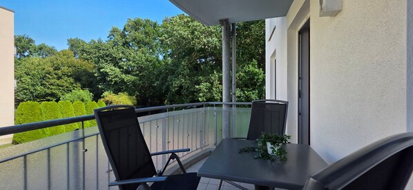 A balcony with two chairs and a table, surrounded by a metal railing, overlooking a green landscape with trees.
