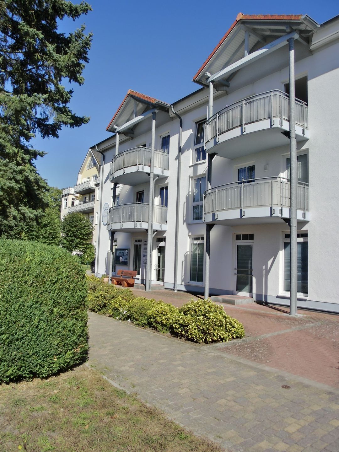 Um edifício residencial de vários andares com varandas, entrada pavimentada e um arbusto bem cuidado.