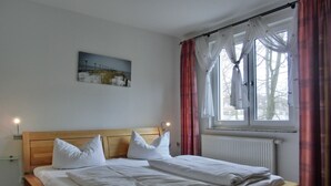 Une chambre avec un lit en bois, une literie blanche, une table de chevet avec une lampe, une fenêtre avec des rideaux et un tableau accroché au mur.