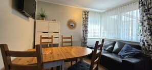 Uma sala de estar com mesa de madeira, duas cadeiras, um sofá de couro preto, TV fixada na parede e janela com cortinas florais.