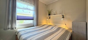 A bedroom with a bed, a window with curtains, a shelf with decorative items, and a radiator.
