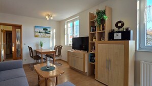 A compact living space with a sofa, dining table, chairs, a TV, and built-in shelving with books and decorative items. - Vacation apartment L41 "Morgensonne" - 3-room apartment (approx. 54 m²) for 4 people with balcony (Graal-Müritz)
